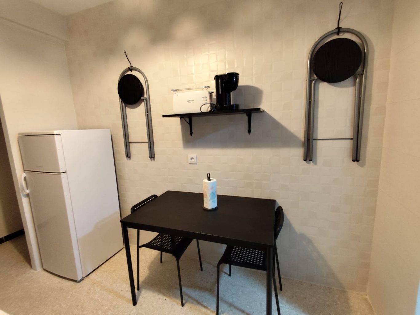 Cocina Cocina con mesa negra, sillas, nevera y estante en la pared.